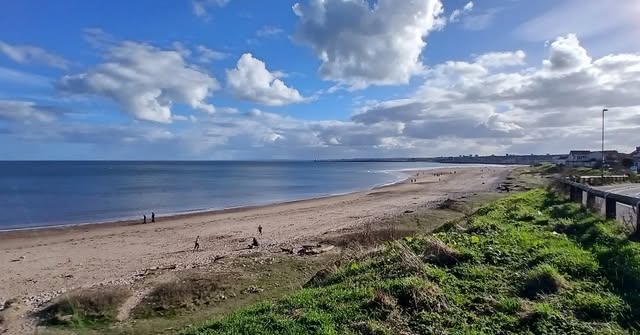 A broad sandy beach and blue sea beneath a sky filled with white clouds, viewed from a grassy bank
